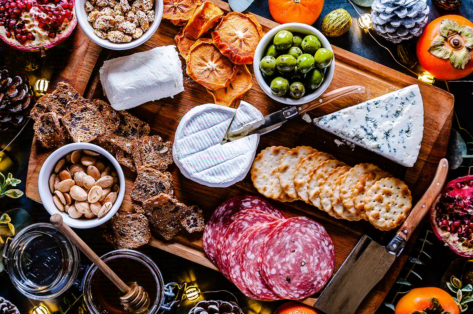 A close up of cheeses, olives, fruits, crackers, nuts and spread on a charcuterie board