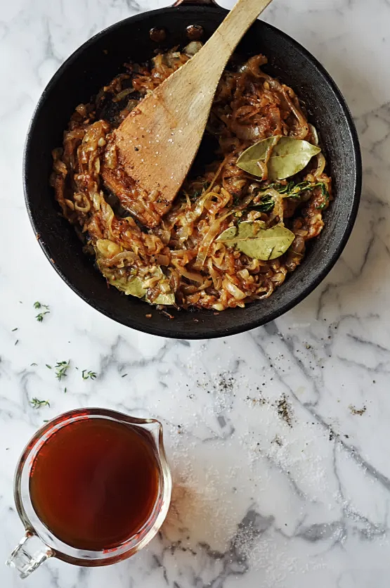 a skillet with beef, onion, and bay leaves sautéing on a table next to a cup of beef stock