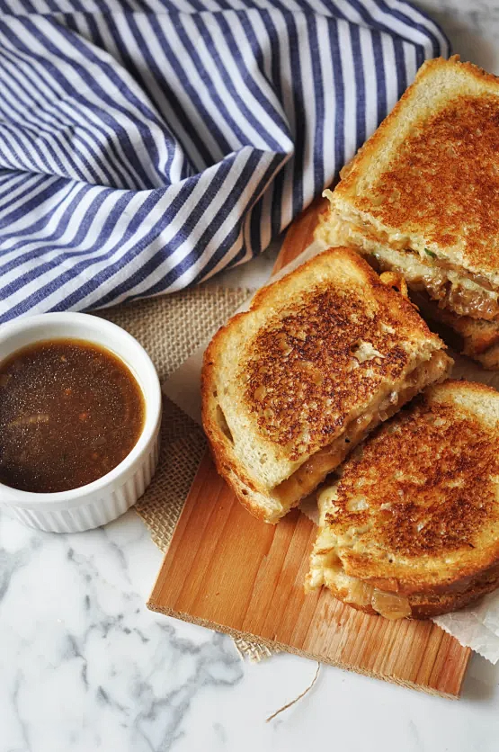 A sliced grilled cheese sandwich in three pieces on a cutting board next to a serving dip and a cooking towel